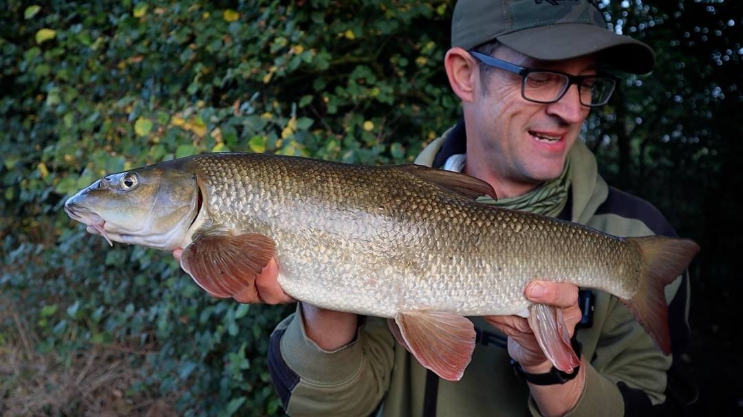 A man wearing a cap and glasses holds a large barbel horizontally in both hands in front of leafy bushes.