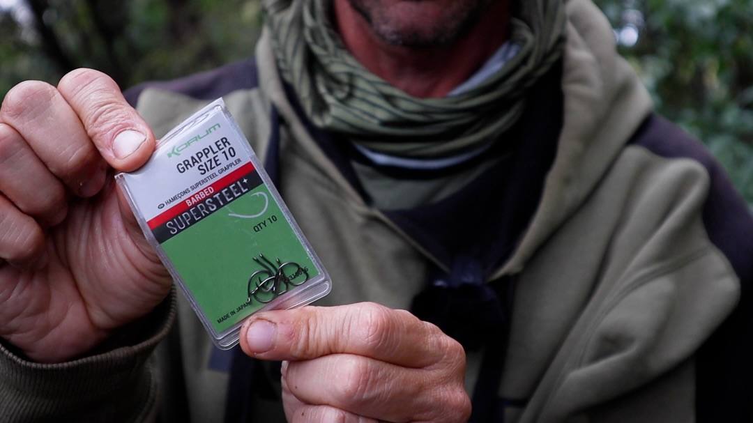 An angler in an olive jacket and neck gaiter holds a clear packet of Korum Grappler size 10 barbed hooks towards the camera.