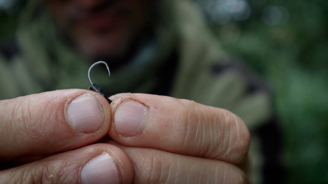 Close-up of an angler holding a curved fishing hook between their fingers, with their face and green clothing blurred in the background.