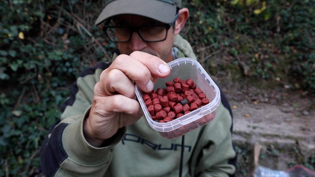 A man wearing a cap, glasses and a green hoodie holds a clear plastic tub of red bait pellets towards the camera outdoors.