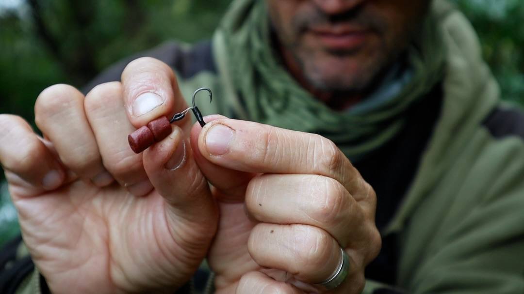 An angler in green clothing holds a fishing hook and hair rig with two reddish-brown cylindrical baits close to the camera.