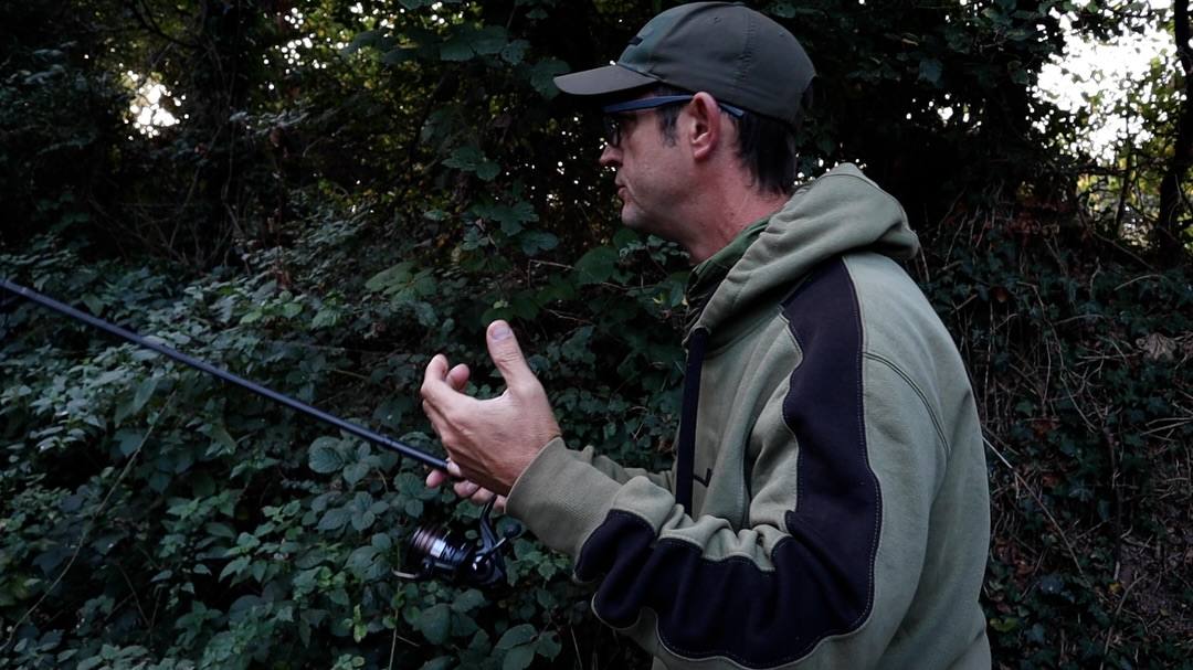 An angler wearing a green cap, glasses and a green and black hoodie holds a fishing rod and reel while gesturing with one hand beside dense foliage.