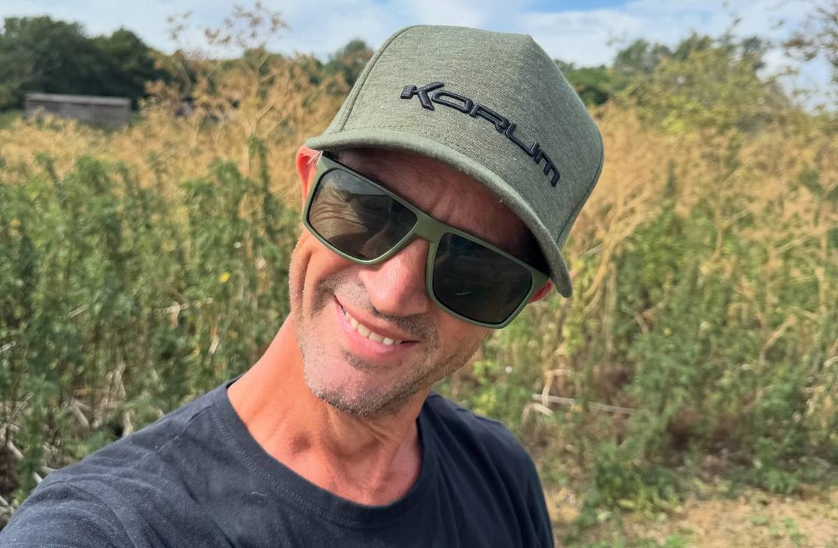 Here It Is author smiling in a green Korum cap and sunglasses beside tall bankside vegetation