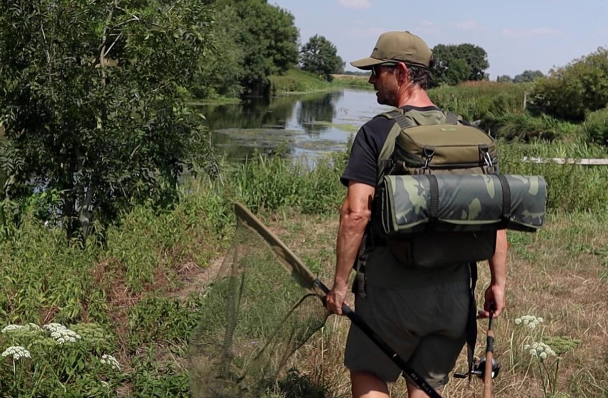 Angler carrying a landing net, rod and rucksack with a rolled mat along a grassy riverbank