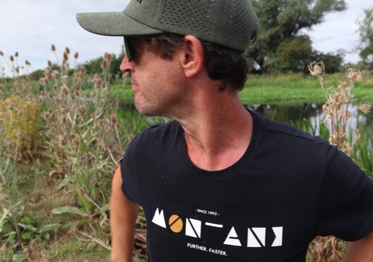 An angler wearing a green cap, sunglasses and a black Montane T-shirt beside the river