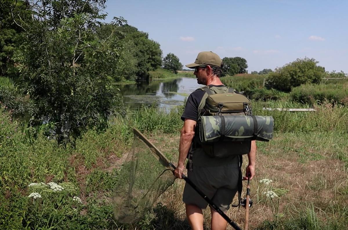 Angler carrying a rucksack, rolled camouflage mat, landing net and rod along a grassy riverbank