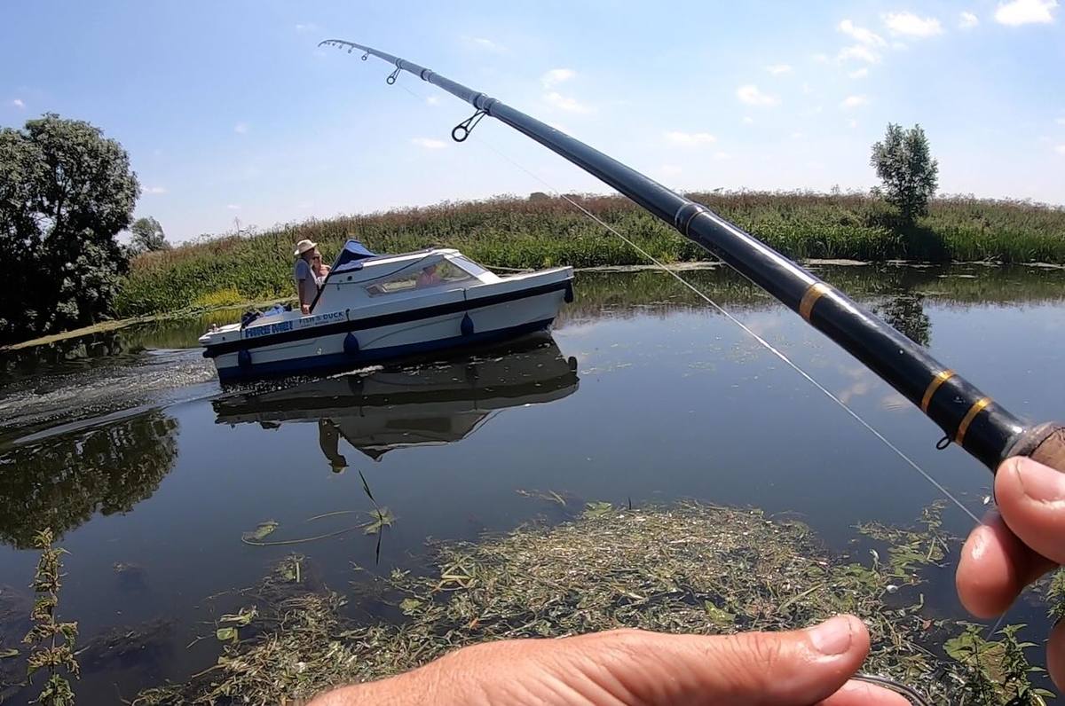 View past an angler’s hands and raised fishing rod as a small cabin boat passes on the river