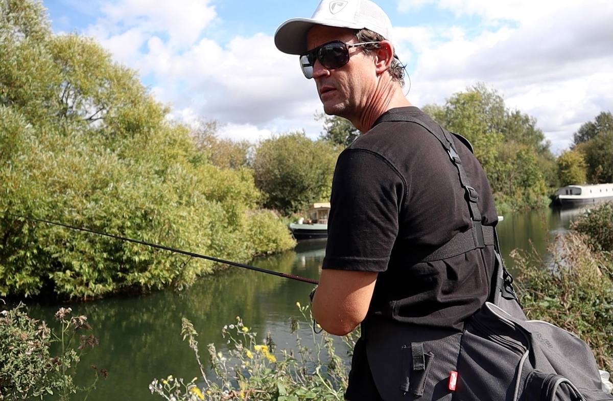 Angler holding a lure rod beside a tree-lined waterway with moored boats