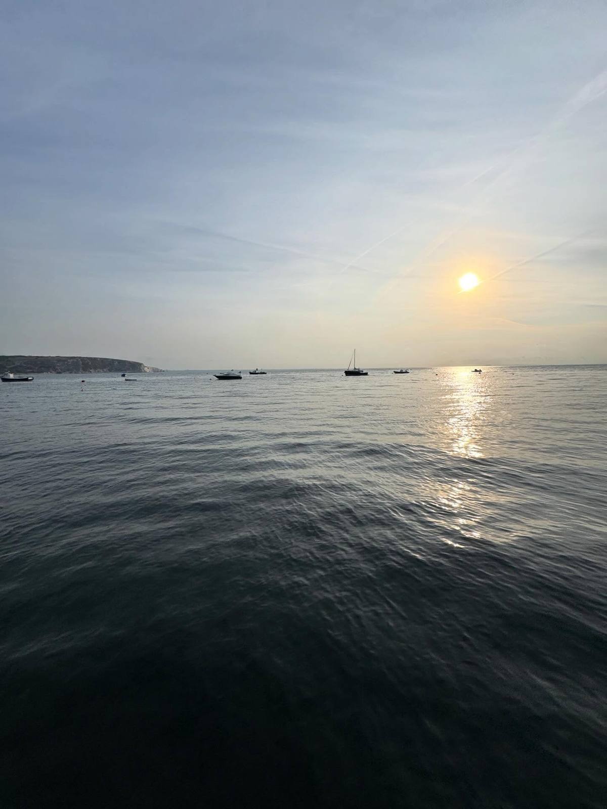 Sun low over the sea with moored boats and a headland in the distance