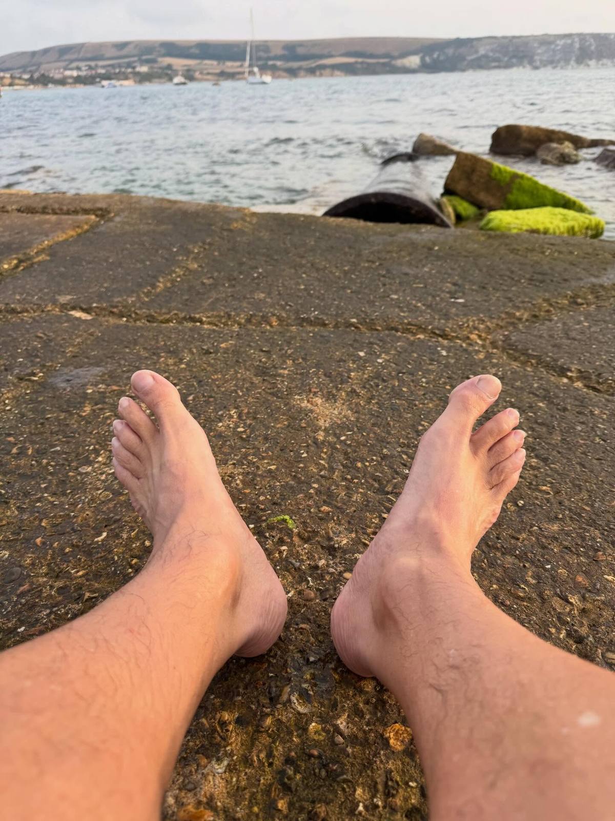 Bare feet resting on the waterfront with boats and coastal hills across the bay