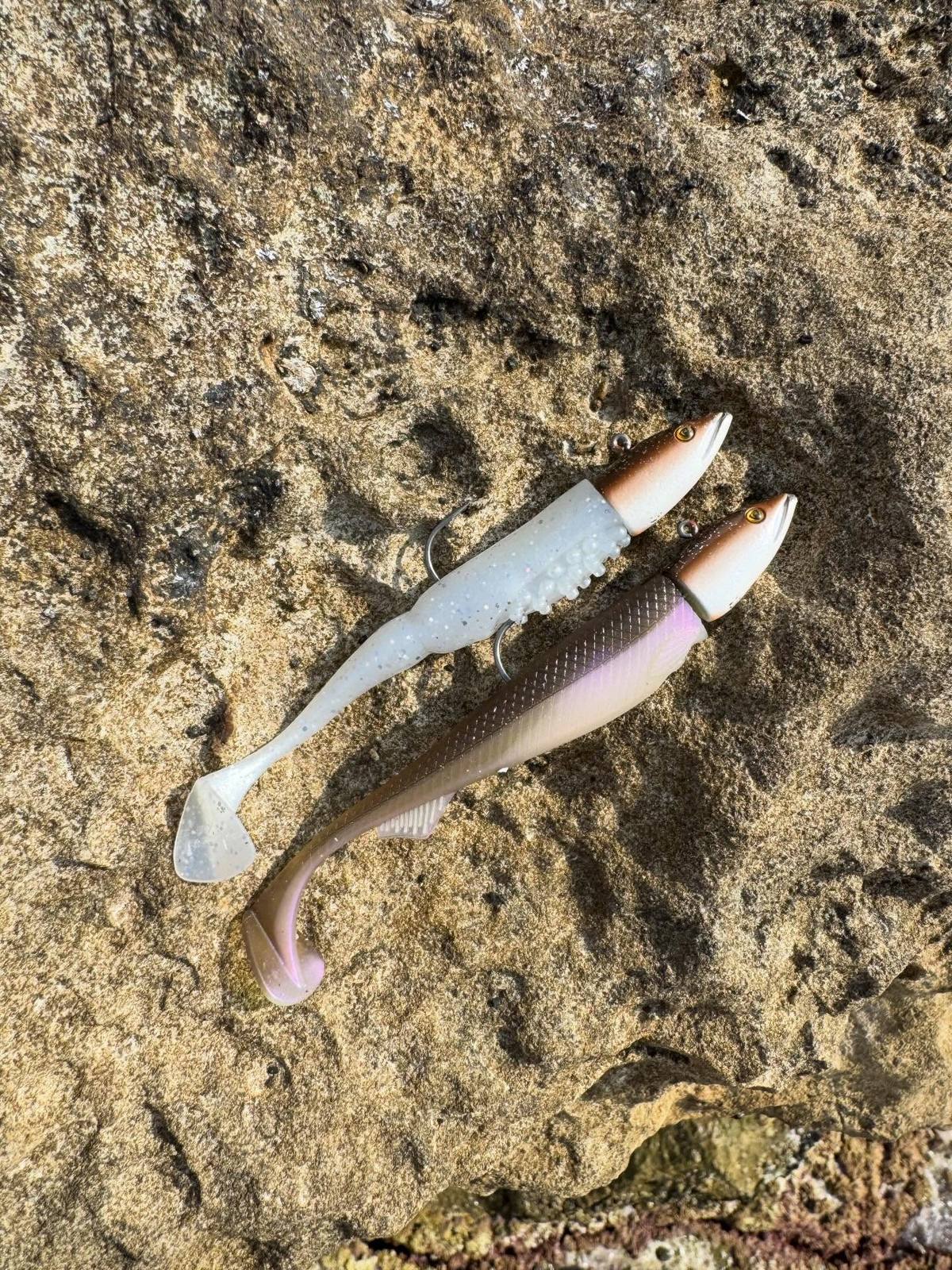 Two paddle-tail soft plastic lures with weighted fish-shaped heads on a rock