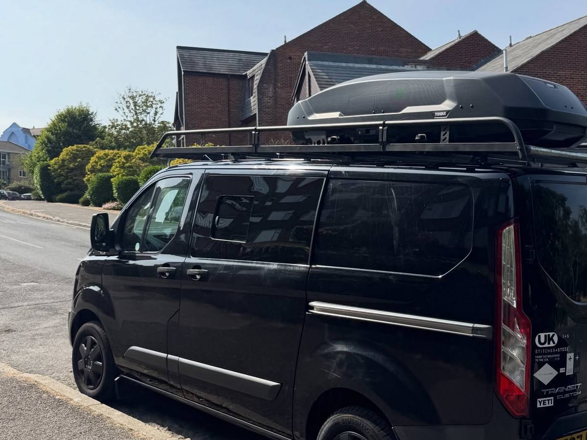 Black Ford Transit Custom van parked on a residential street with a roof rack and roof box