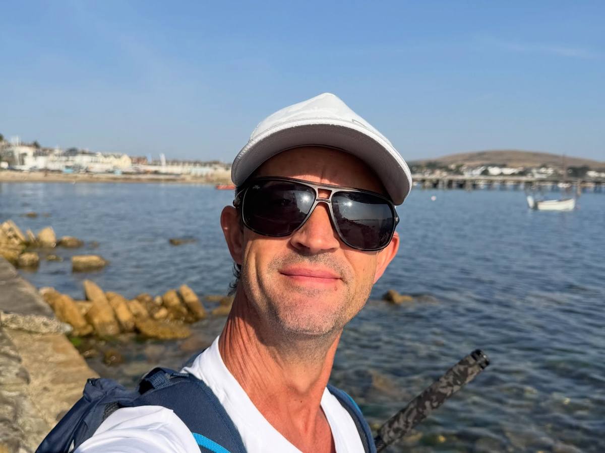 Angler wearing a pale cap and sunglasses beside the sea with boats and waterfront buildings behind him