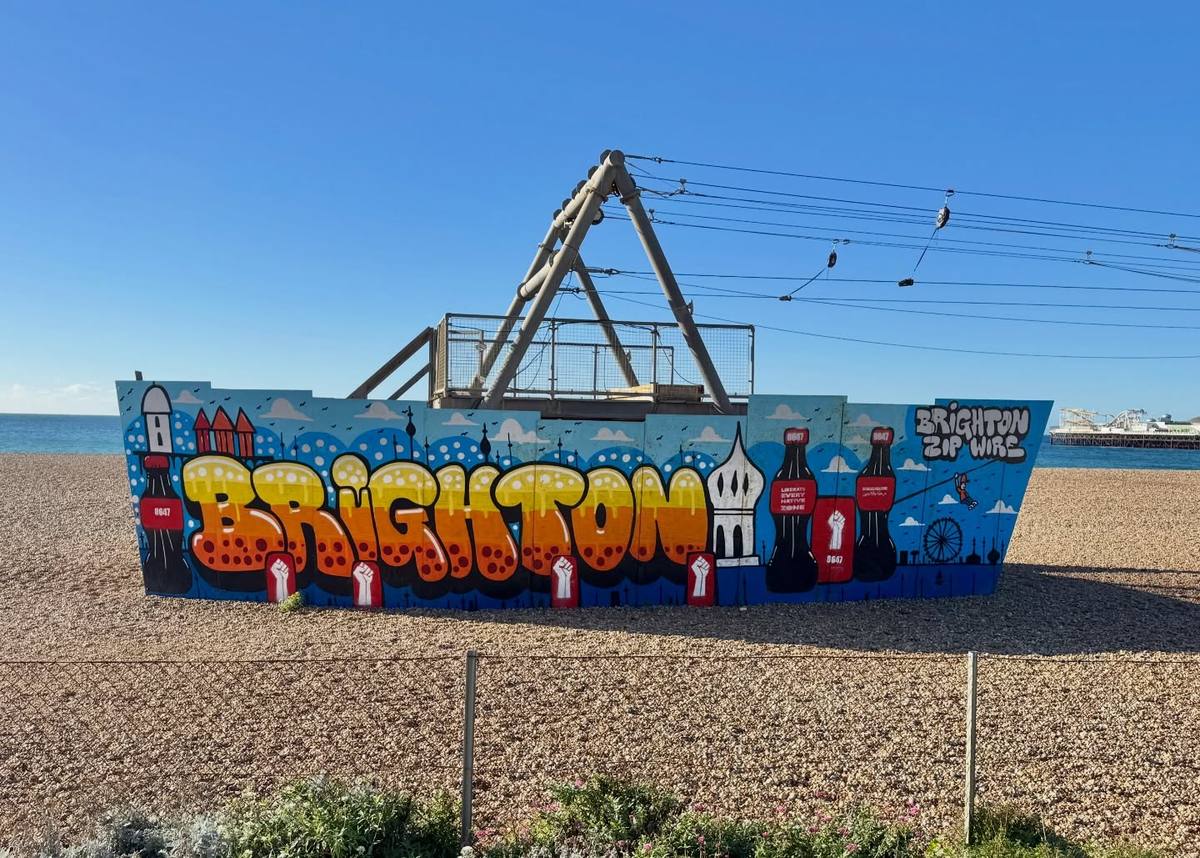 A colourful Brighton mural surrounds a zip wire structure on a shingle beach, with the sea and a pier behind it beneath a clear blue sky.
