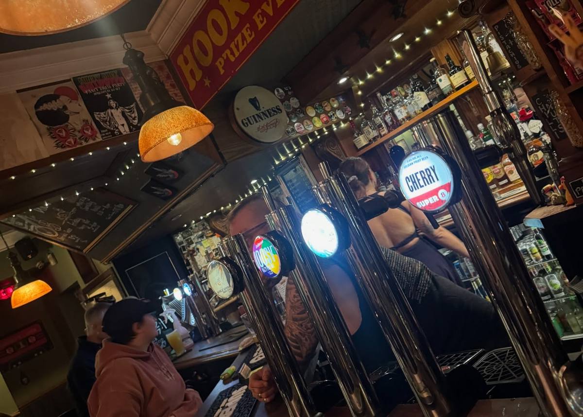 A tilted view across a pub bar shows shiny beer taps, people at the counter, shelves of bottles, hanging lamps and strings of fairy lights.