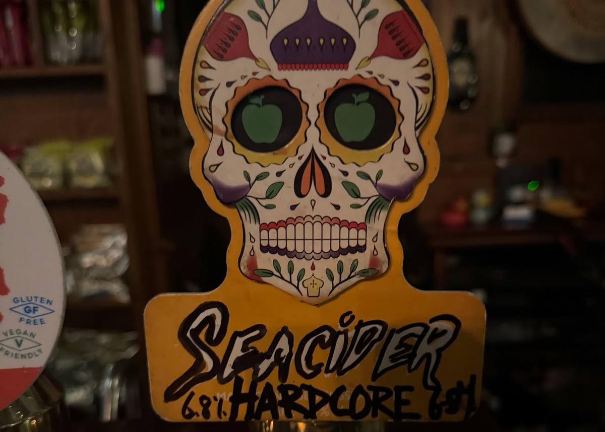 A yellow Seacider Hardcore pump badge features a colourful decorative skull with green apples in its eye sockets, with bottles and glasses blurred behind it.