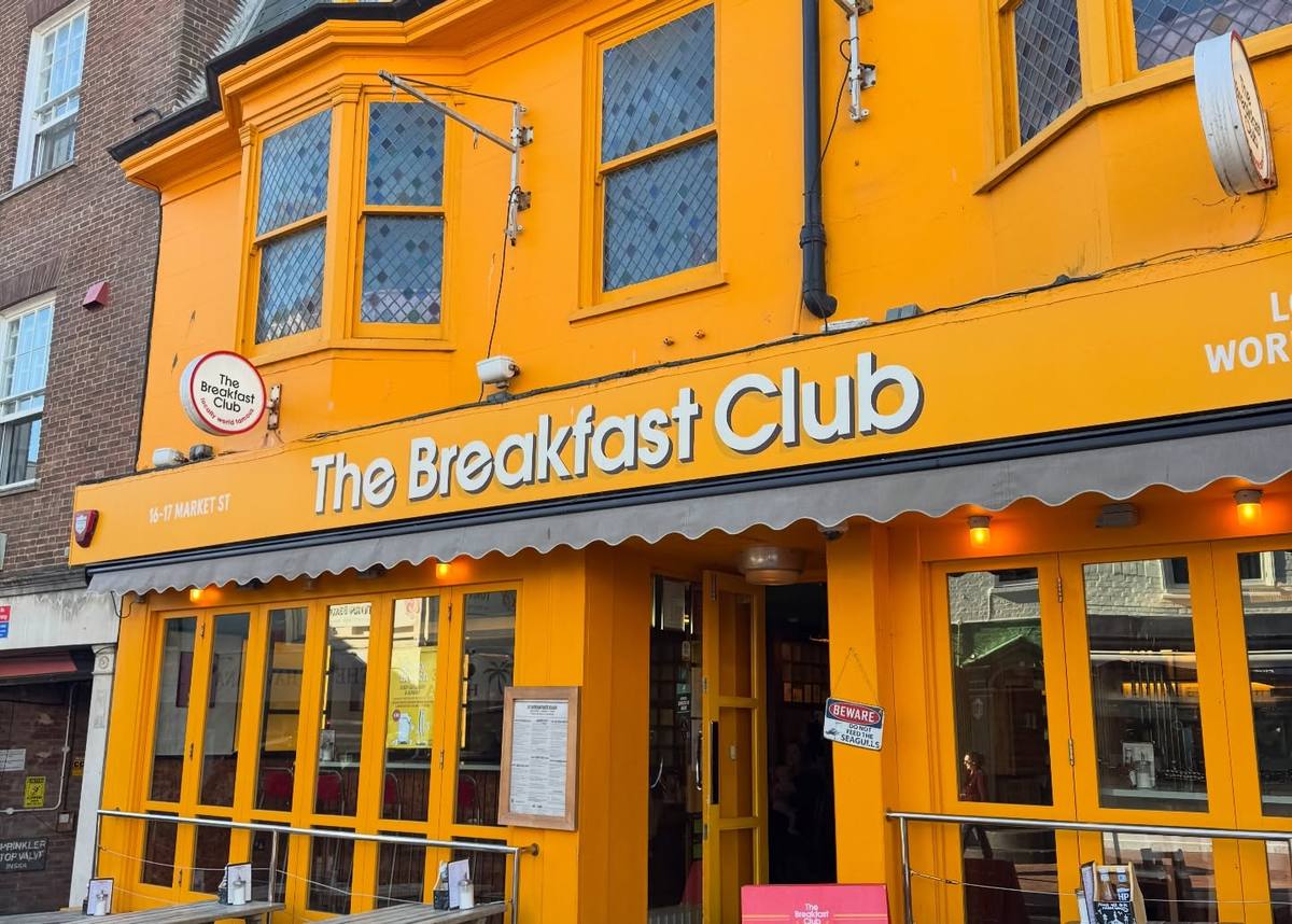 The bright yellow-orange frontage of The Breakfast Club has large white lettering, a grey awning, an open entrance and outdoor tables.