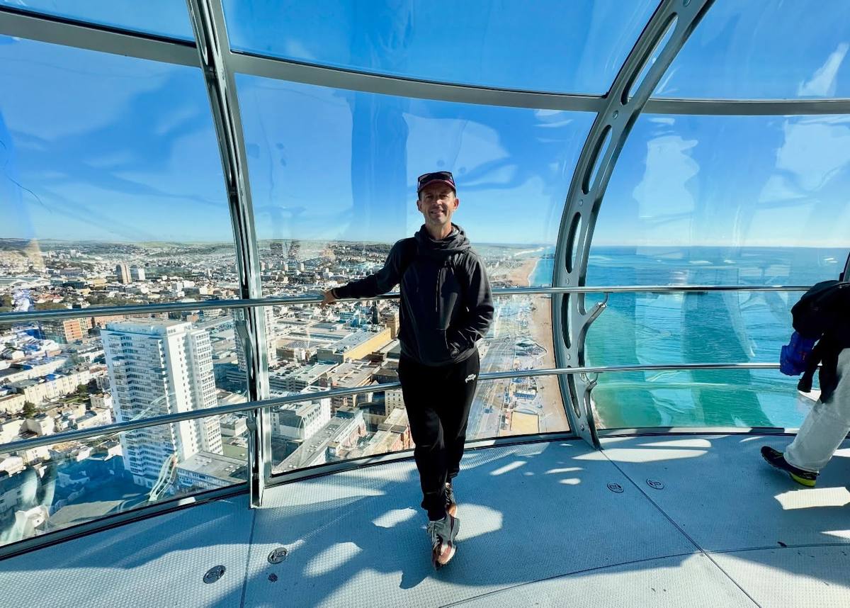 A smiling man in a black hoodie and cap stands beside the railing inside a glass observation pod, high above a coastal city, beach and blue sea.