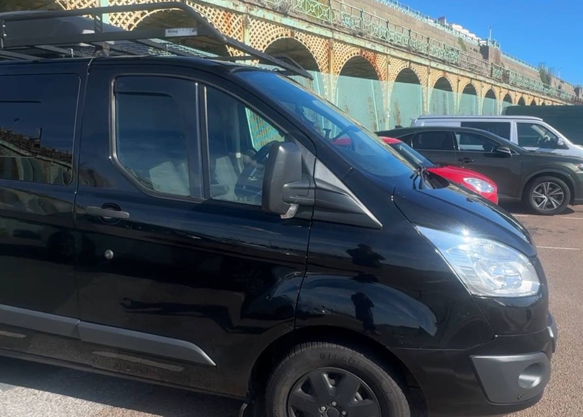 A black van with a roof rack is parked beside other vehicles and ornate metal arches under a blue sky in this image for The Adventure Starvation Is Over.