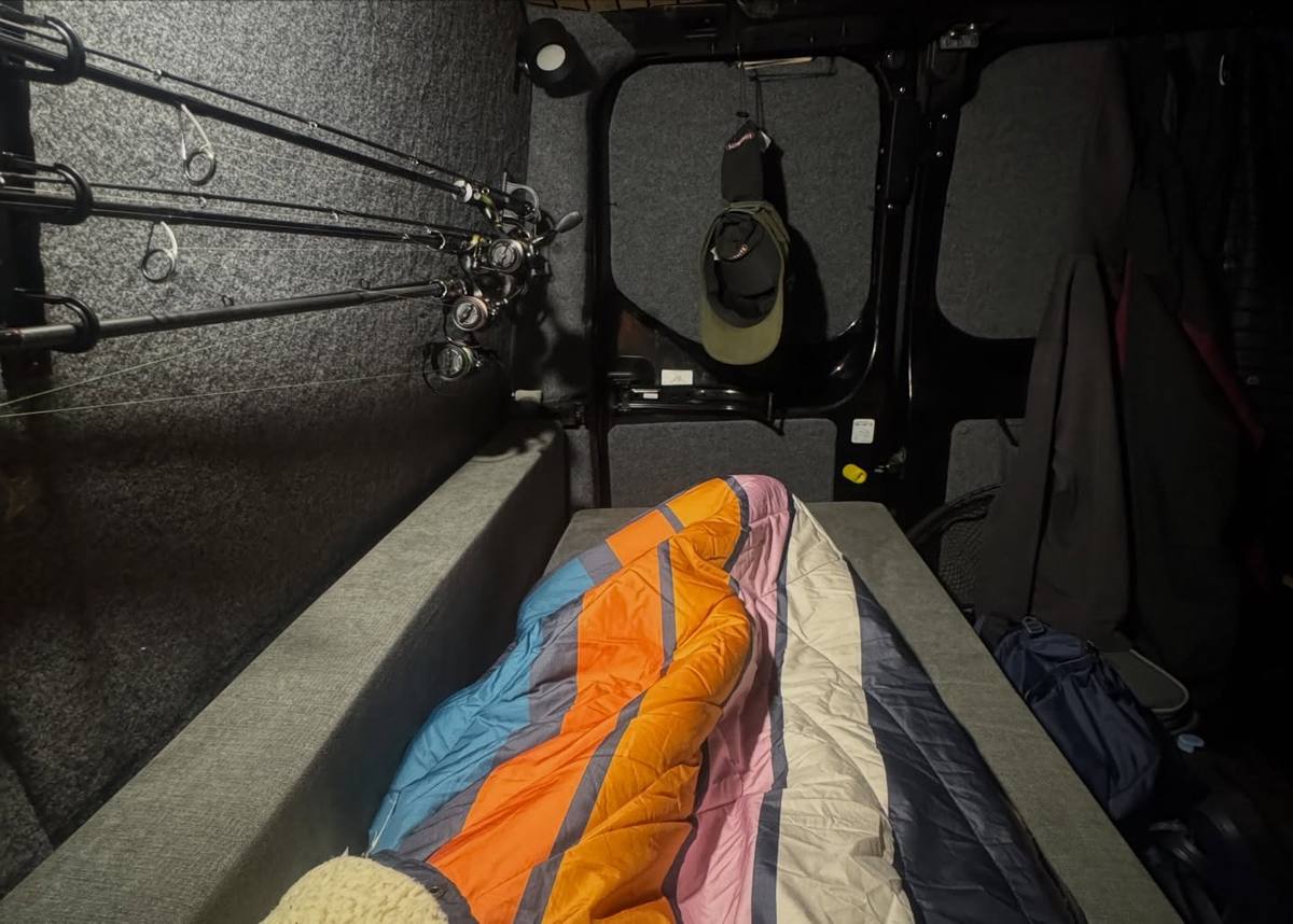Inside a grey-lined van, fishing rods and reels are mounted along the wall beside a bed with colourful striped bedding, with caps hanging on the rear door.
