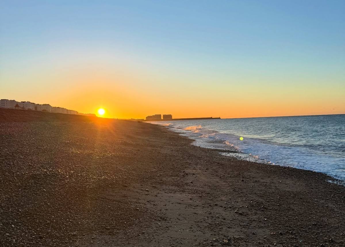A low sun casts golden light across a pebble beach, with waves breaking along the shore, seafront buildings to the left and a breakwater in the distance.