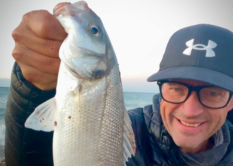 A smiling man wearing glasses, a dark cap and a padded jacket holds a large silver fish close to the camera with the sea behind him.