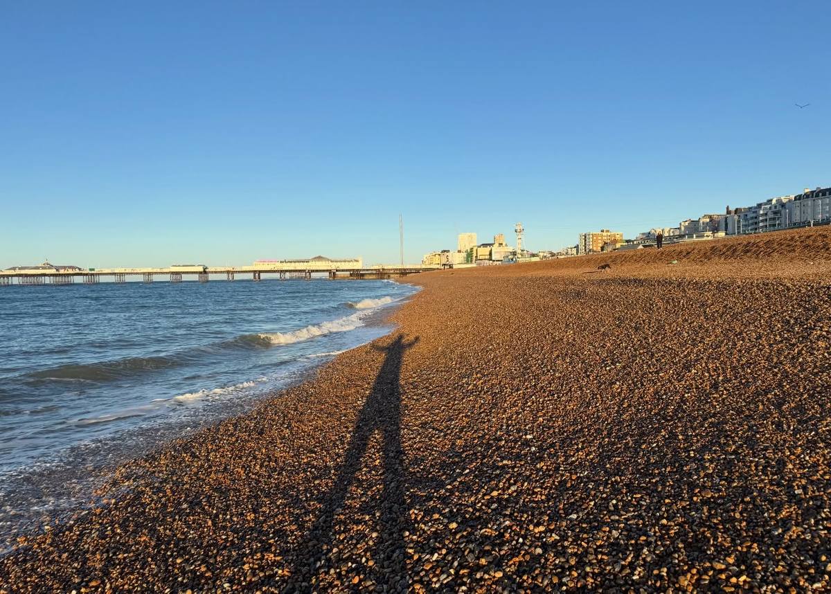 Small waves wash onto a sunlit pebble beach, with a long human shadow in the foreground, a pier to the left and seafront buildings beneath a clear blue sky.