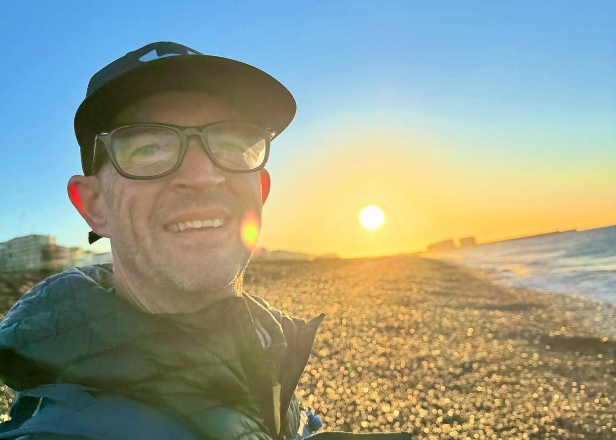 A smiling man wearing glasses, a dark cap and a padded jacket takes a selfie on a shingle beach, with the sea and low golden sun behind him.