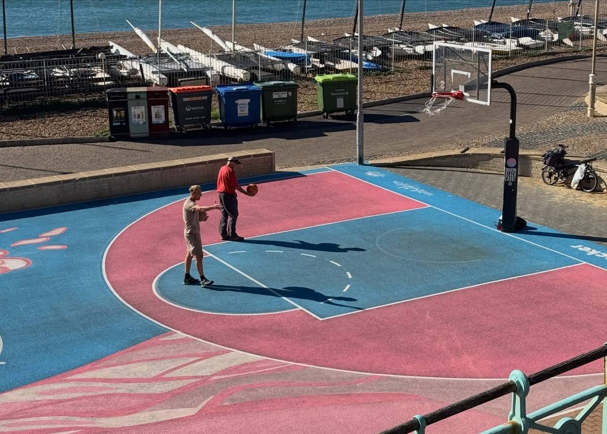 Two people stand on a blue and pink seaside basketball court, one holding a basketball, with bins, sailing boats and the sea behind them.