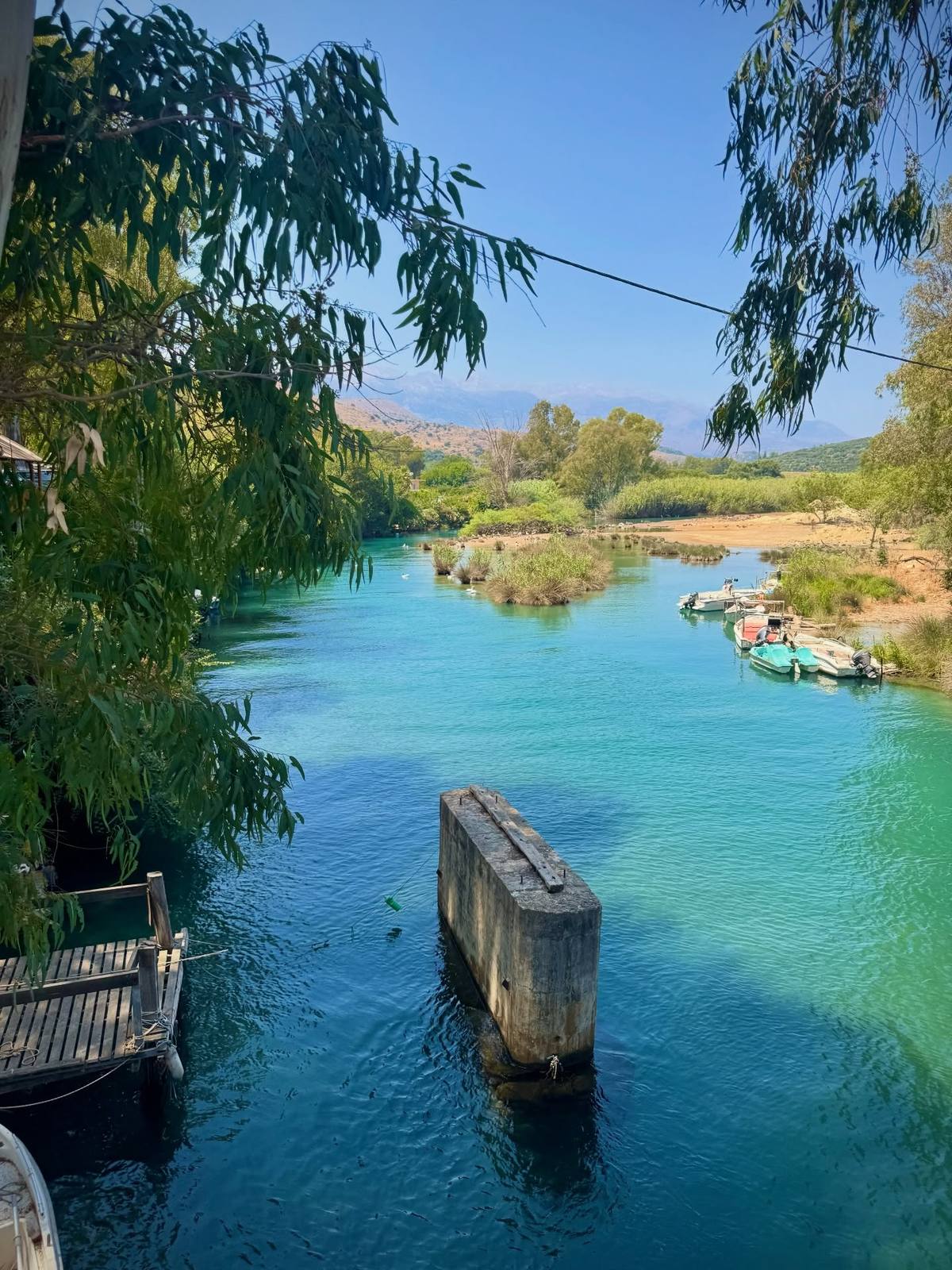 Turquoise water surrounds a concrete pillar beside a wooden landing, with small boats moored along a reedy bank, overhanging branches and distant hills beneath a blue sky.