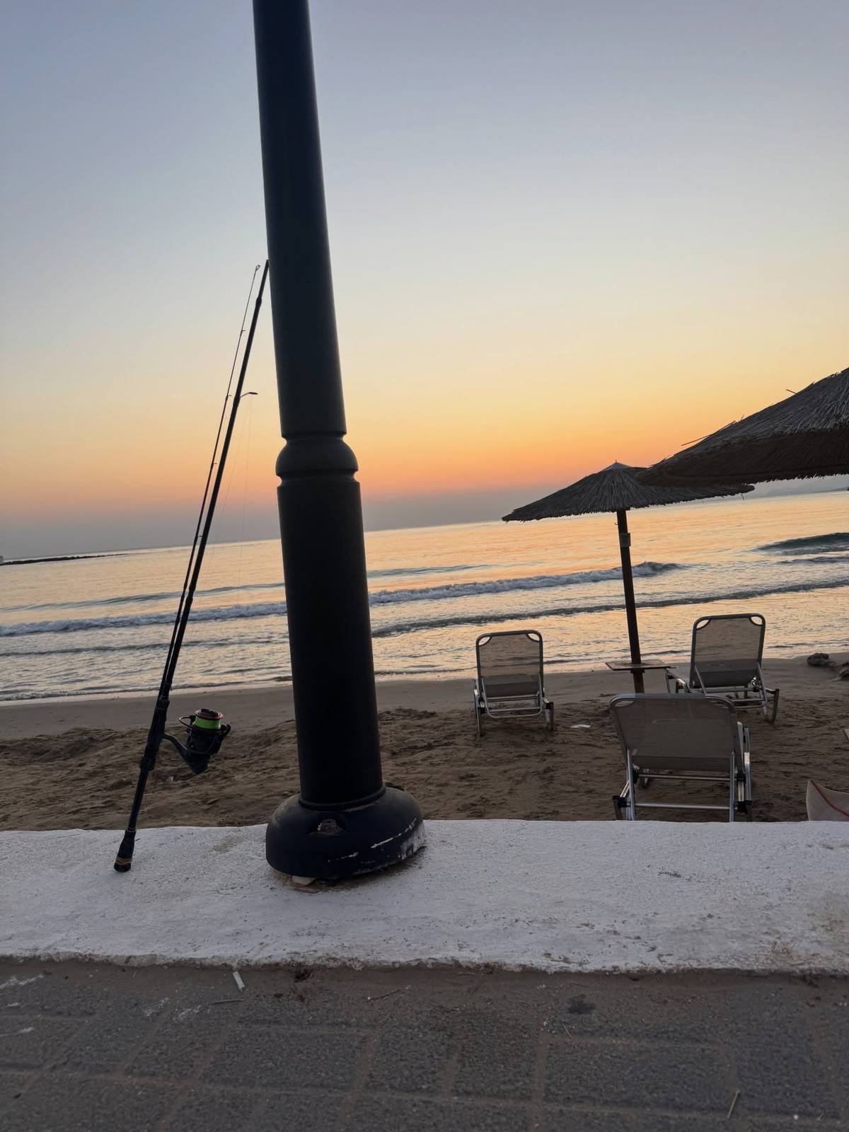 A fishing rod and reel lean against a black post beside a sandy beach with sun loungers, thatched parasols and gentle waves beneath an orange sky, setting the scene for The Dusky Grouper.