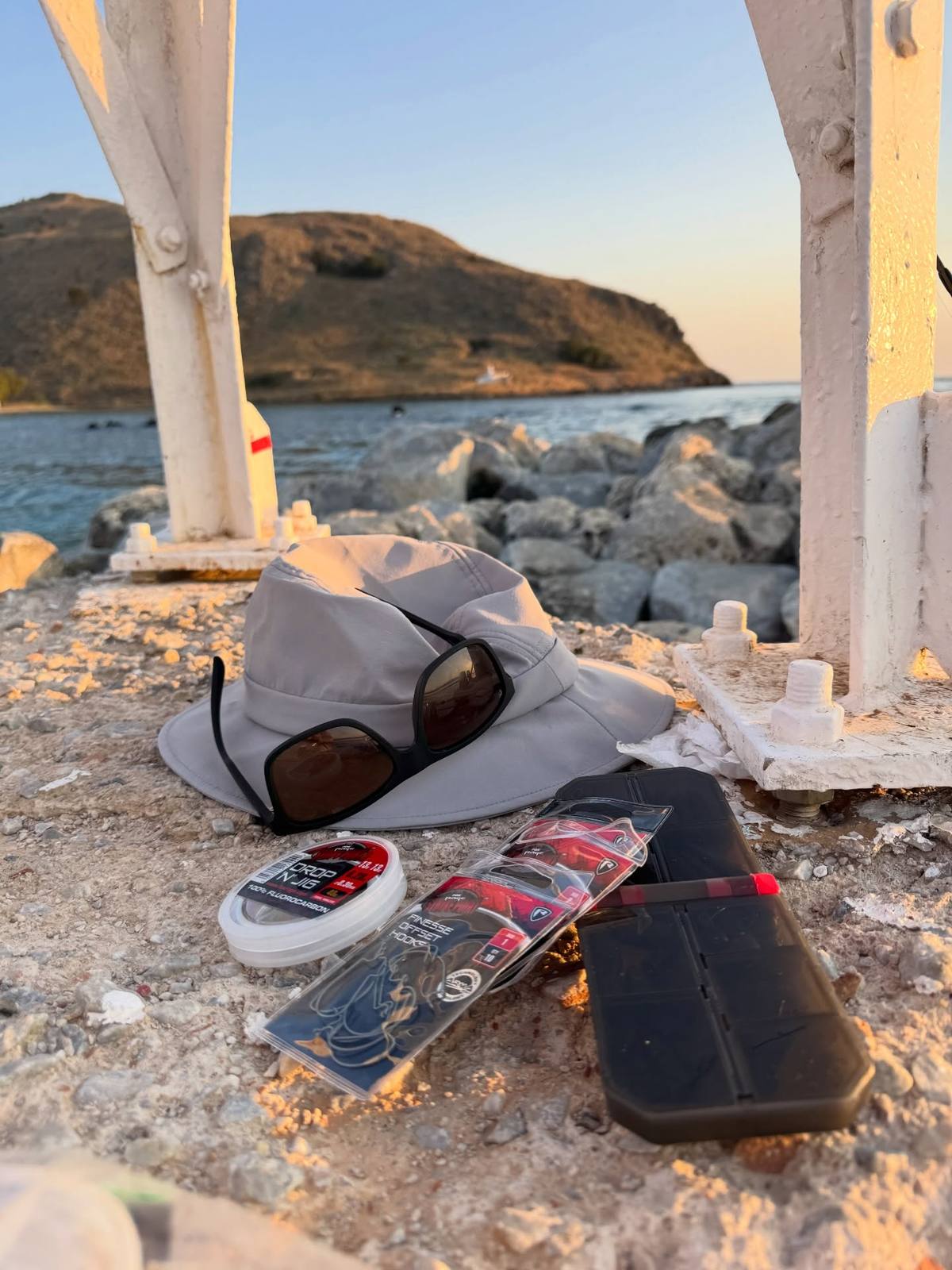 A grey sun hat, sunglasses, fishing line, packets of hooks and a black tackle box rest on a rough stone surface beside white metal supports, overlooking a rocky bay.