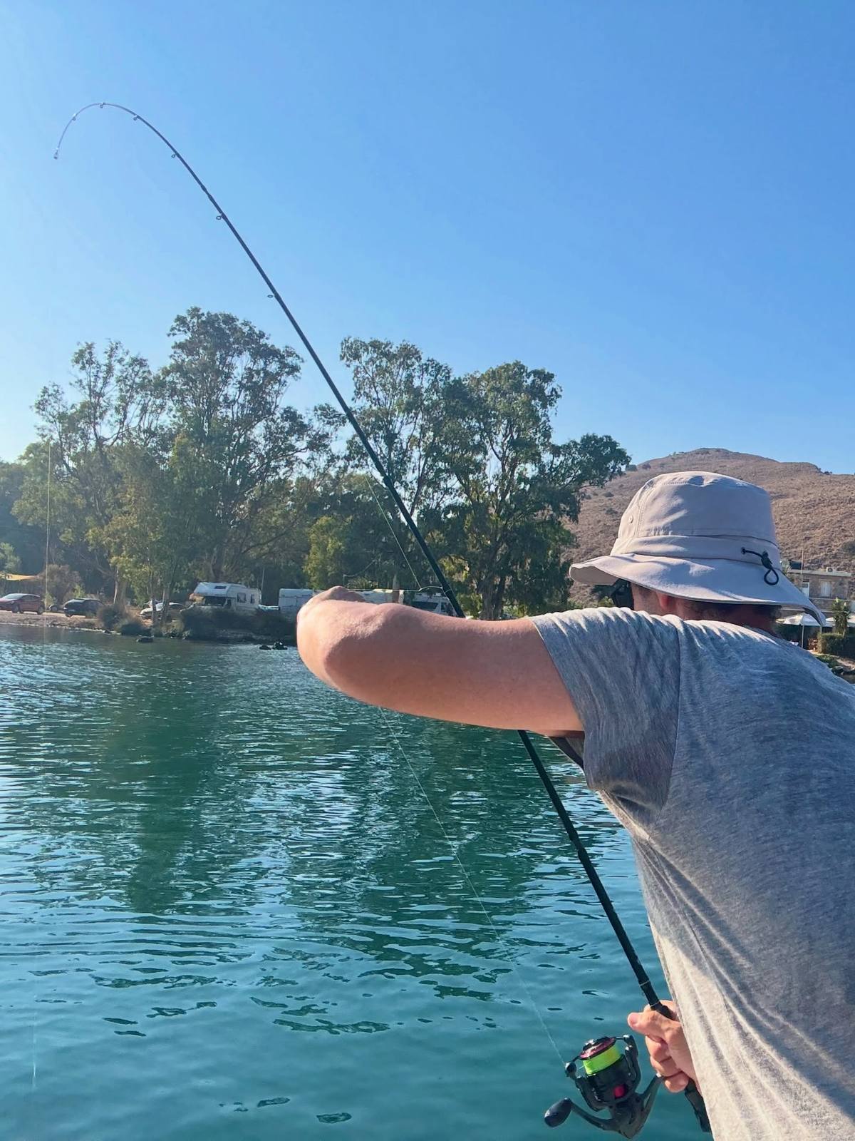 An angler in a pale sun hat and grey T-shirt holds a bent fishing rod over blue-green water, with trees, vehicles and dry hills along the opposite shore.