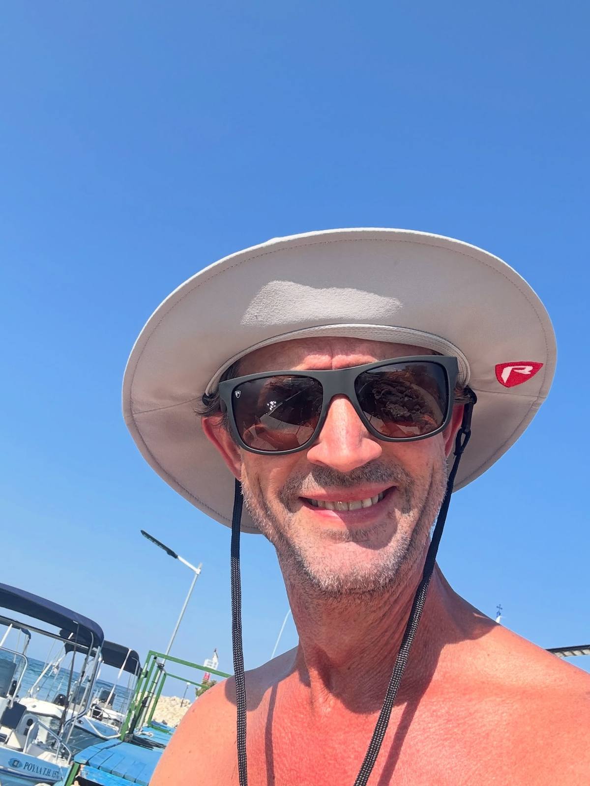 A smiling, bare-chested man wears dark sunglasses and a wide-brimmed beige hat, with boats and blue sky behind him.